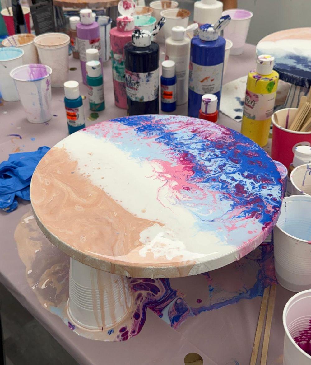 Colorful acrylic pour painting in peach, white, pink and blue on a round canvas surrounded by paint bottles