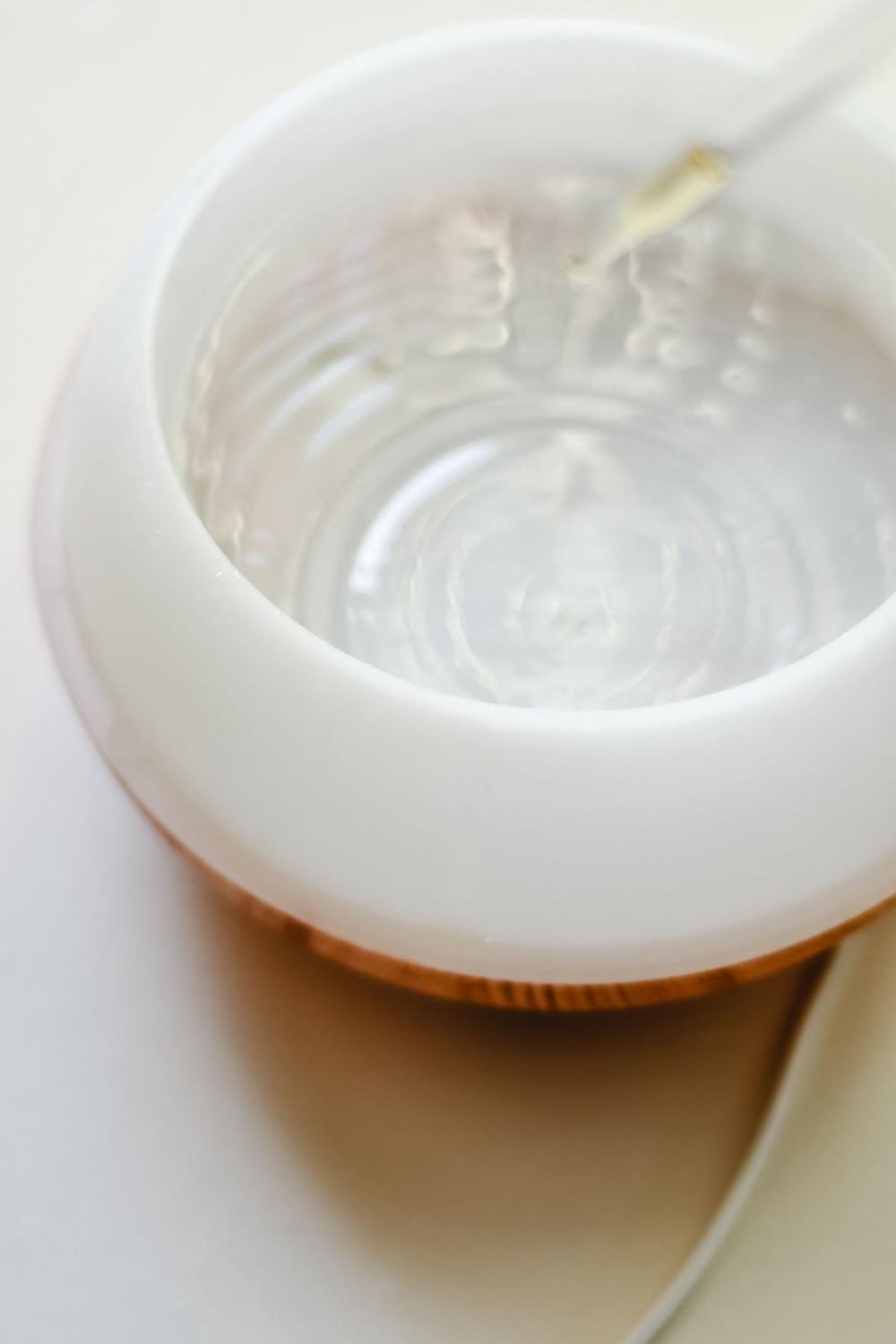 An open white diffuser with a wooden base as drops of essential oil are added to the water