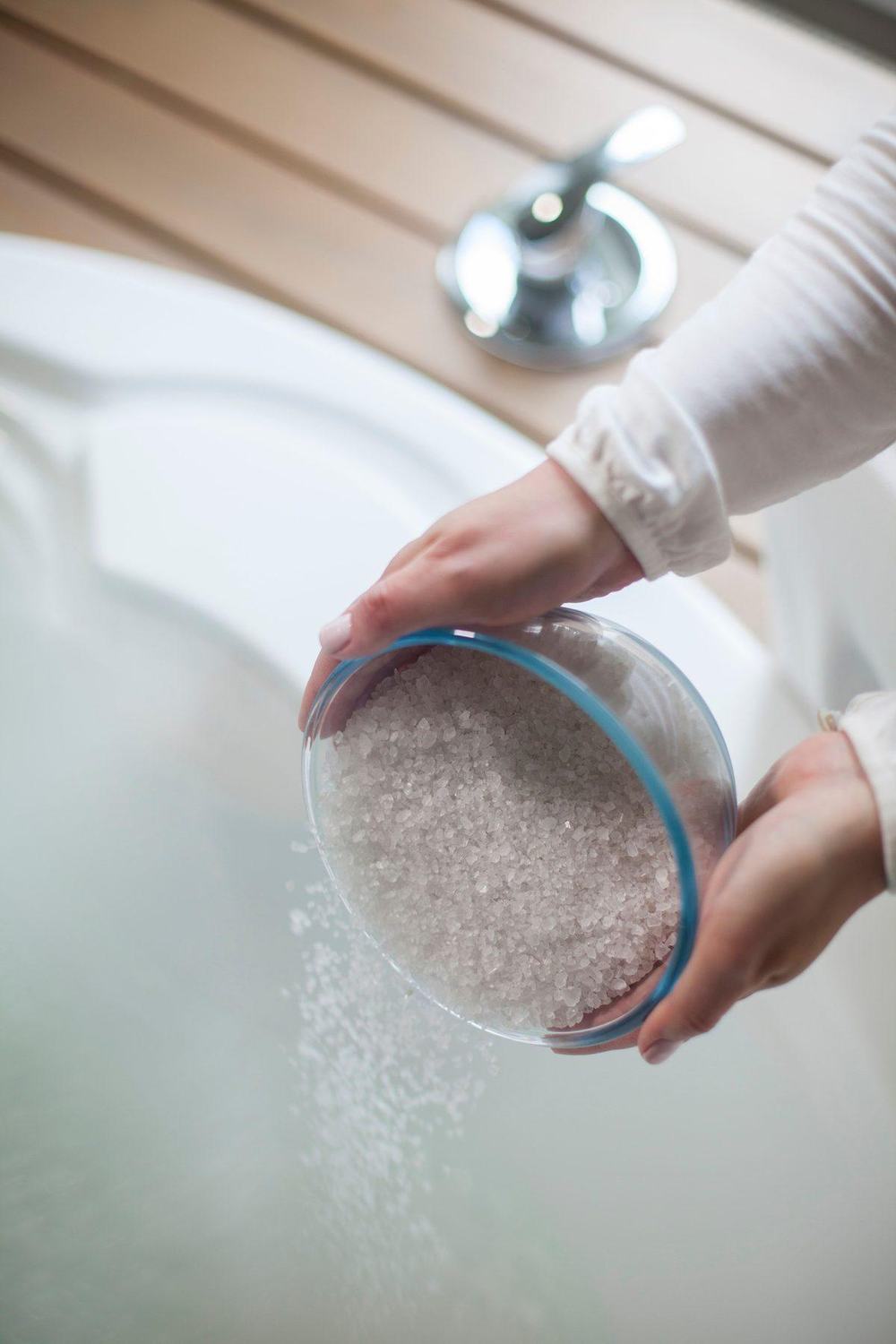 Hands pouring coarse bath salt into a filled white bathtub