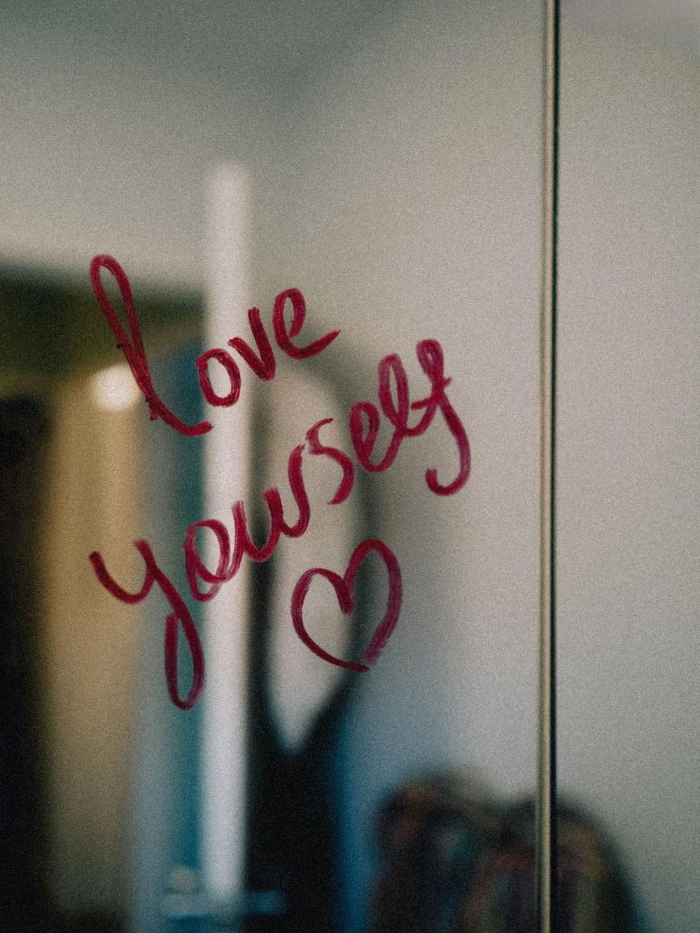 The words love yourself and a heart written in red lipstick on a mirror