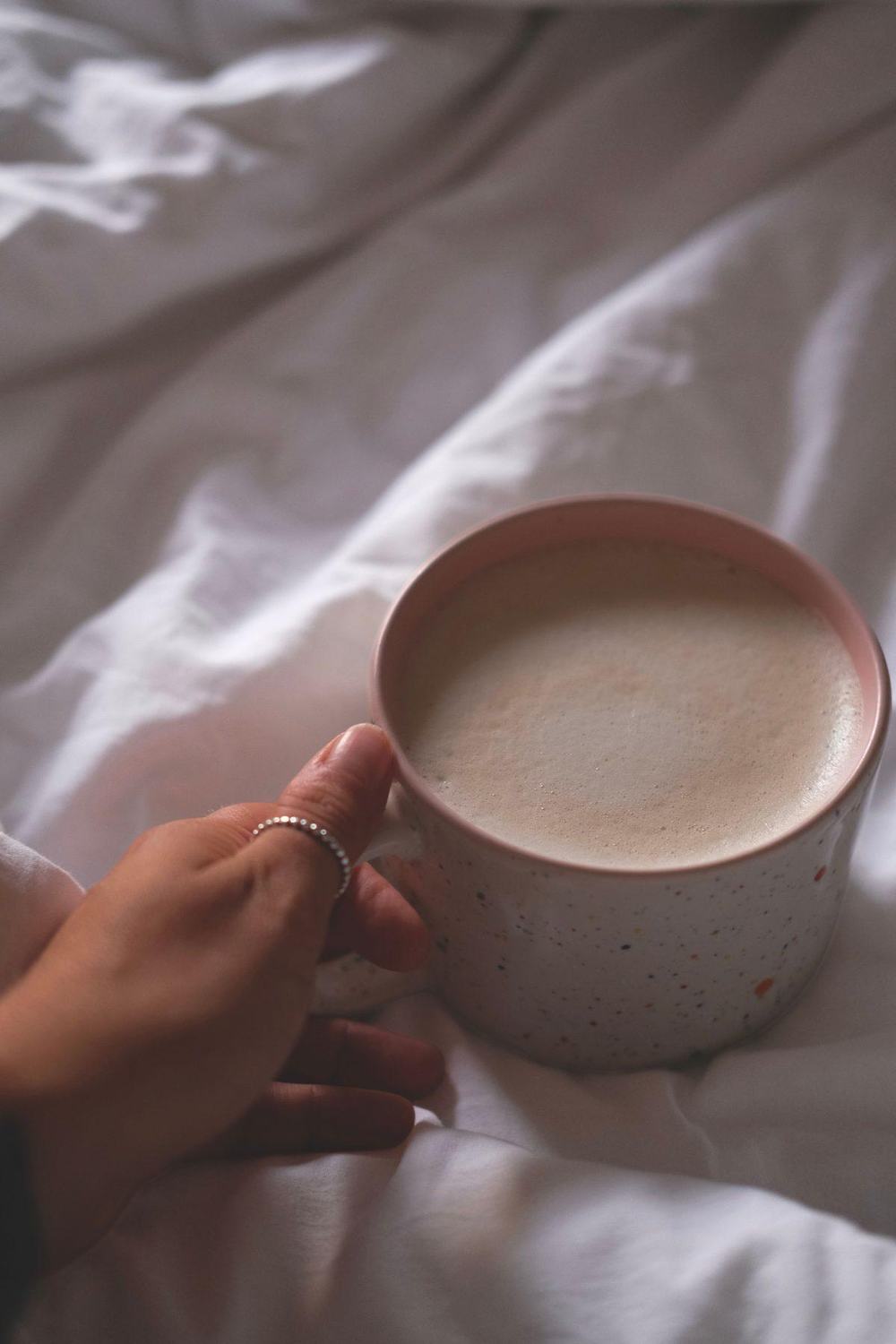 Hand holding a speckled handmade ceramic mug with a pink rim on white bedding