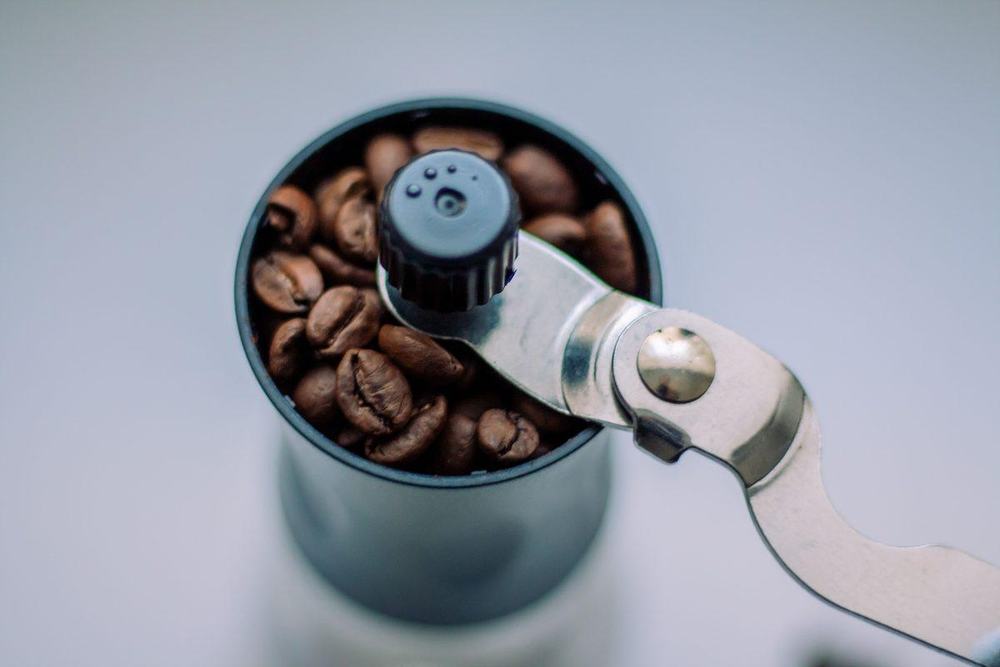 Top view of a manual coffee grinder filled with roasted coffee beans and a steel handle