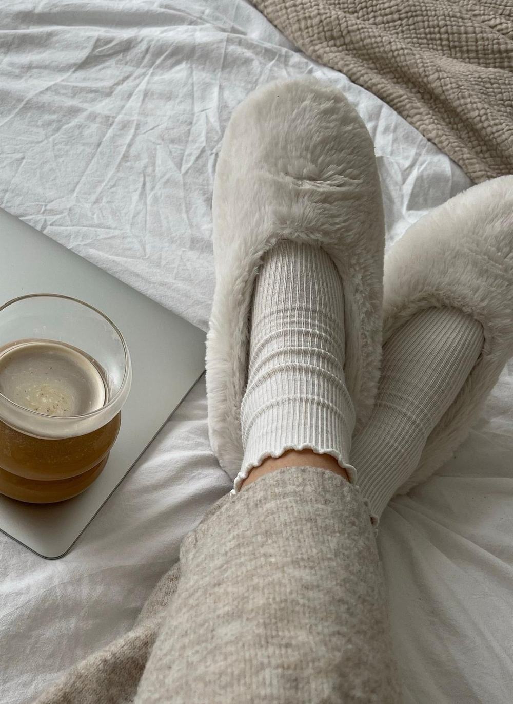 Soft cream faux fur slippers and ribbed socks on a bed with a cup of coffee