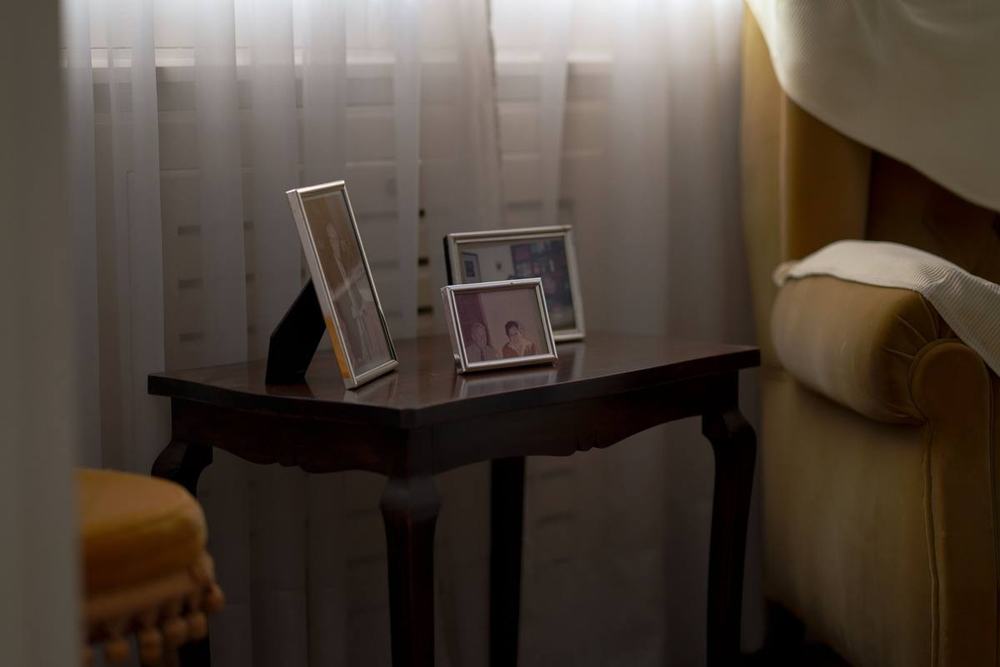 Framed family photos on a dark wooden side table by a window with sheer curtains