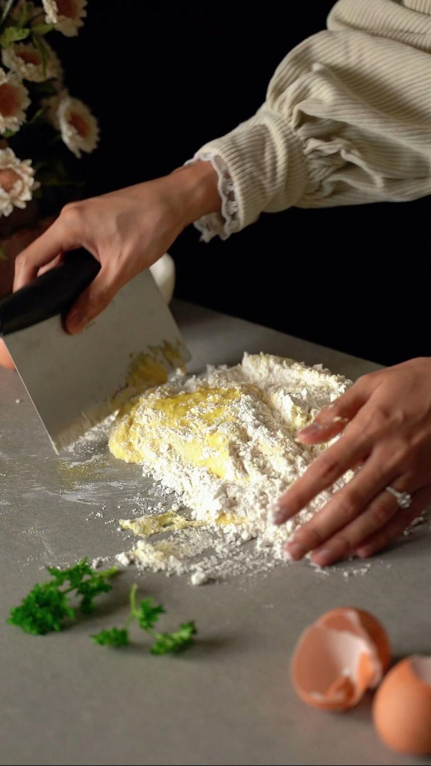 Hands mixing flour and eggs on a grey counter with a metal dough scraper
