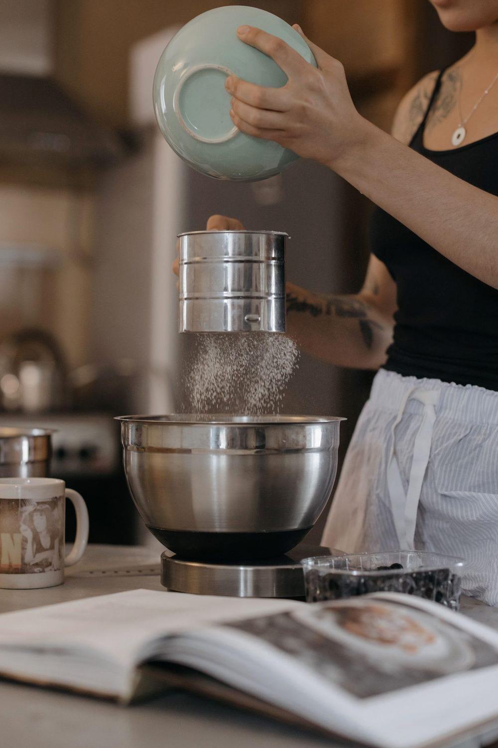 Hand holding a stainless steel flour sifter as flour falls into a mixing bowl