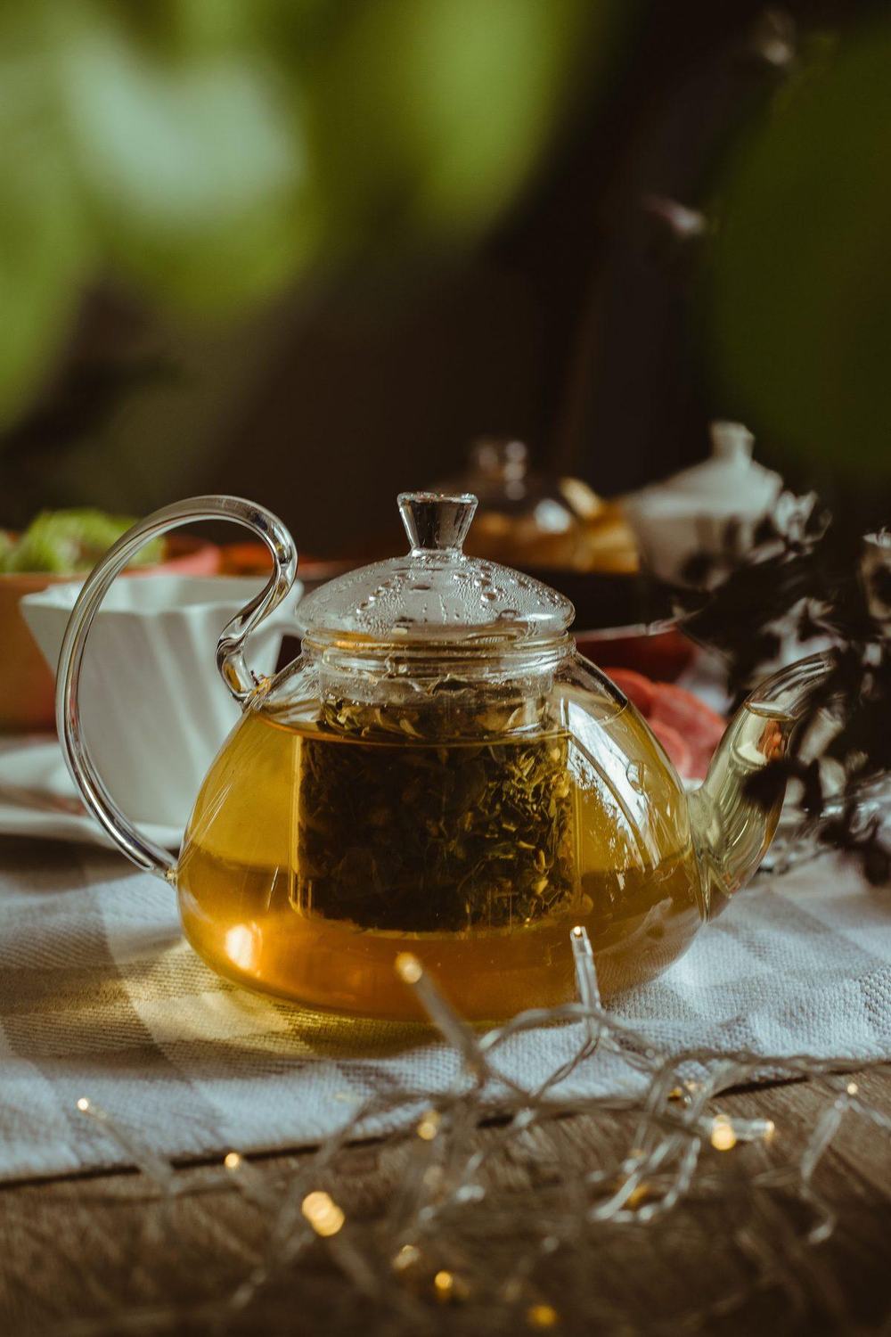 Clear glass teapot with loose leaf tea in an infuser on a linen tablecloth with fairy lights