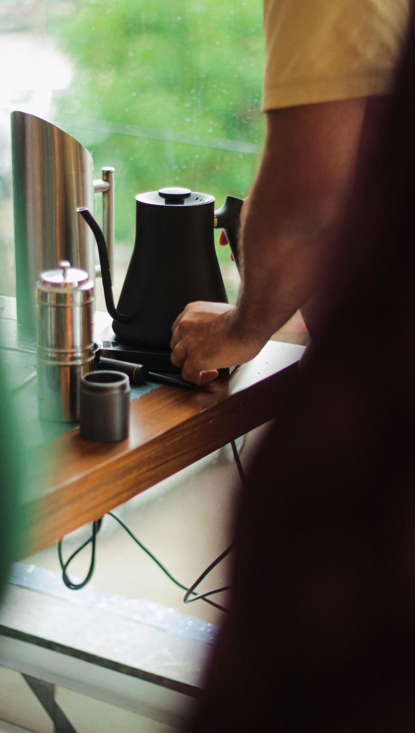 A matte black gooseneck kettle on a wooden counter by a rainy window