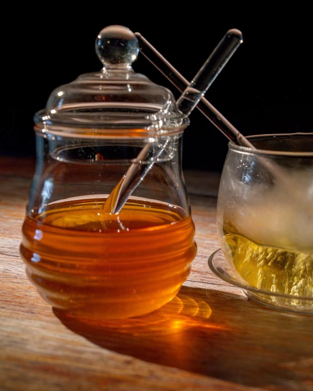 A glass honey jar with a lid and glass dipper next to a cup of tea on a wooden table
