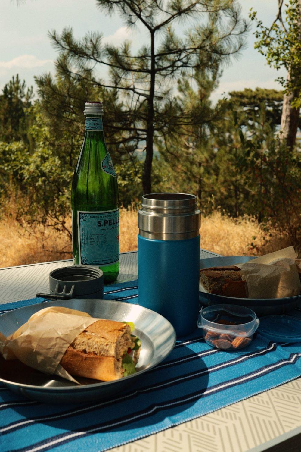 Blue insulated food jar thermos on a picnic table with sandwiches among pine trees