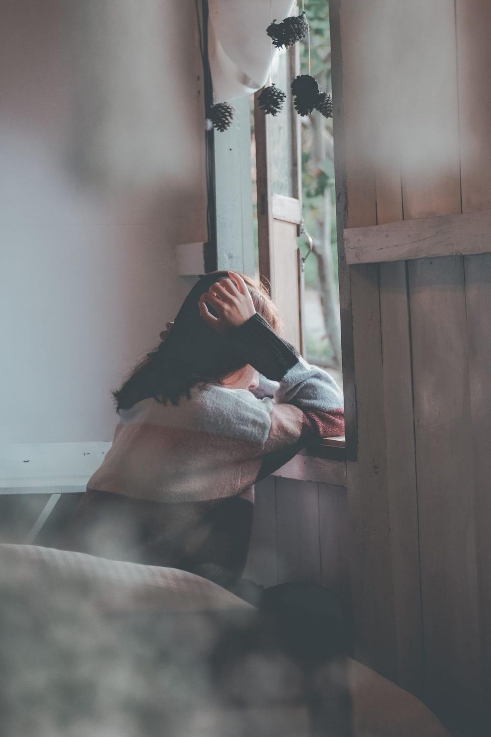 A tired woman leaning on a windowsill with her hand on her head, face hidden