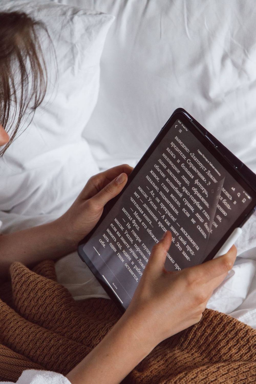 A woman reading in bed under a knit blanket, holding a reading device