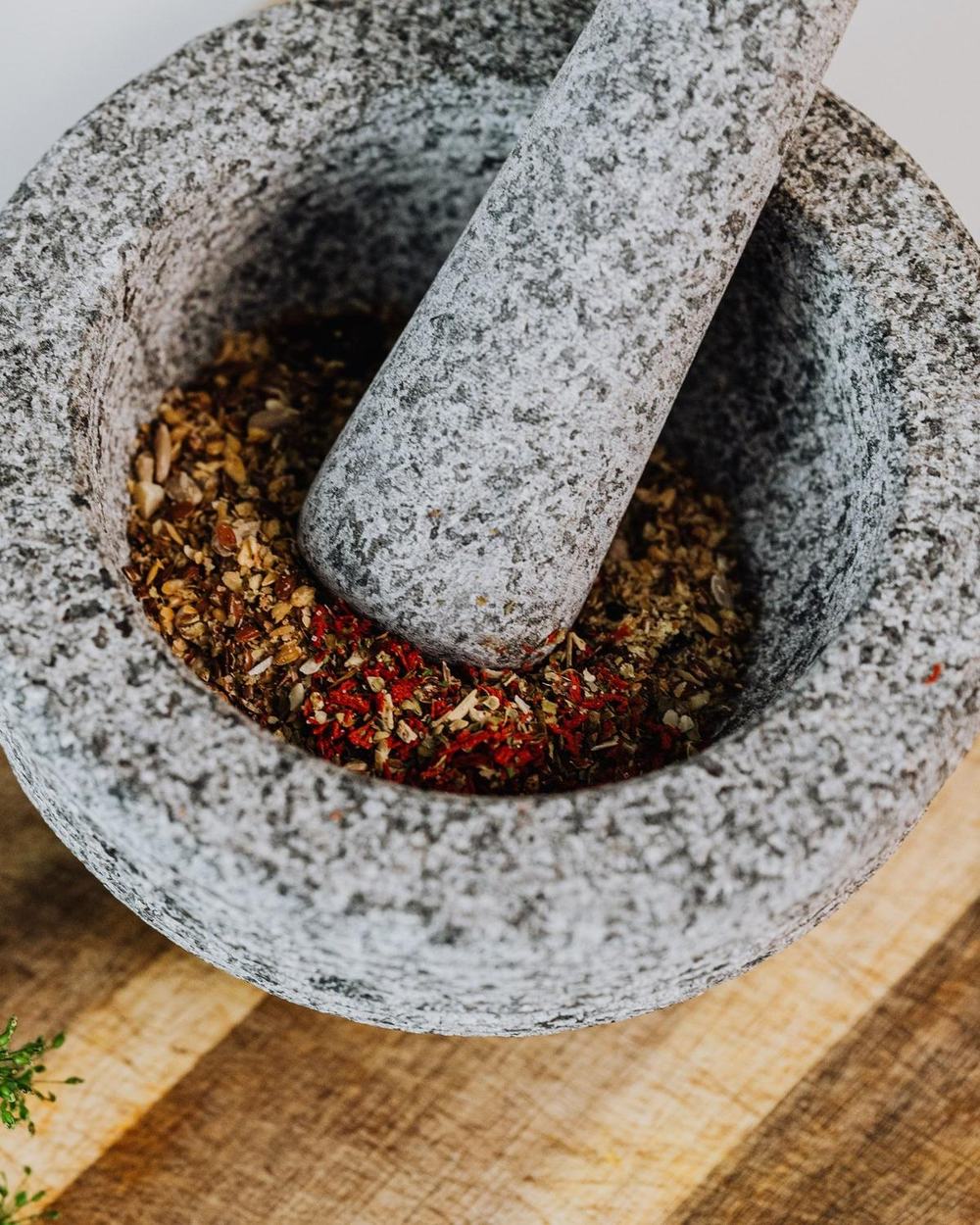 A grey granite mortar and pestle with crushed spices and chili on a wooden board