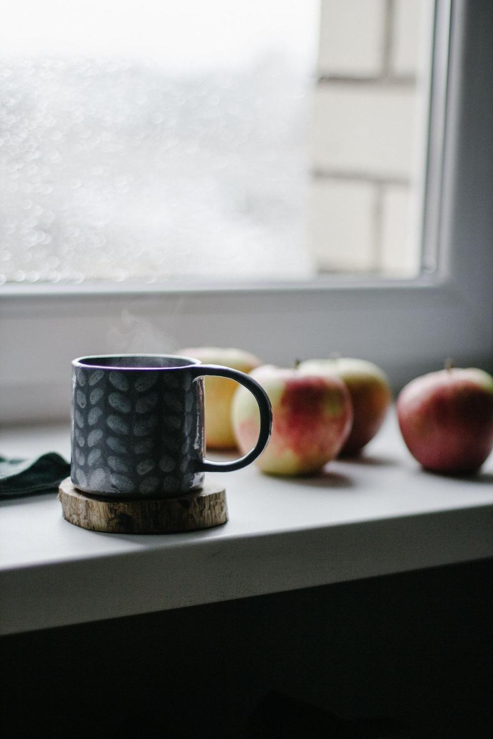 A steaming mug on a wooden coaster by a misty window with red apples
