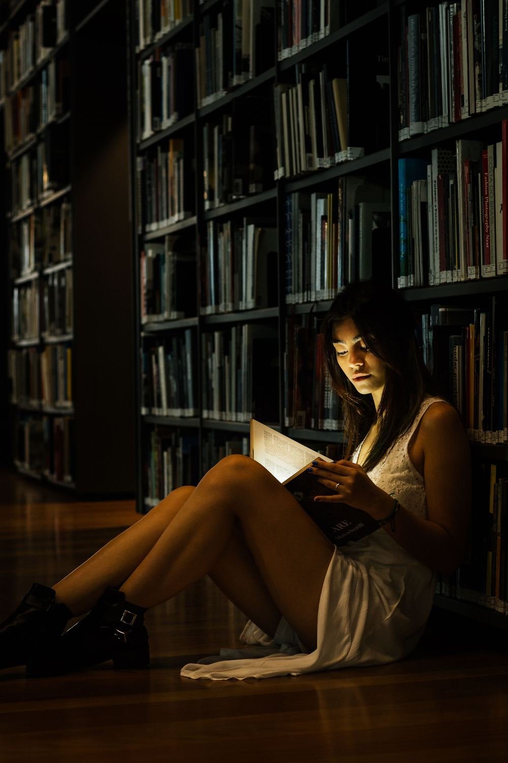 A woman sitting on the floor of a dark library reading a book in a pool of warm light