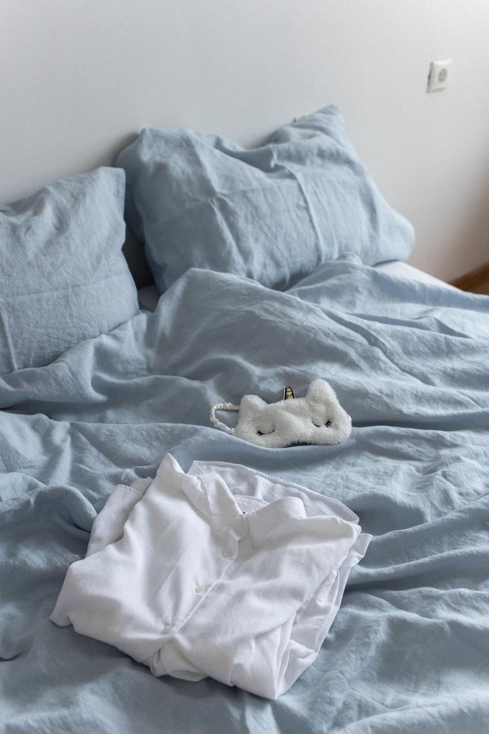 A folded white cotton pajama shirt on a soft blue linen bed with a sleep mask