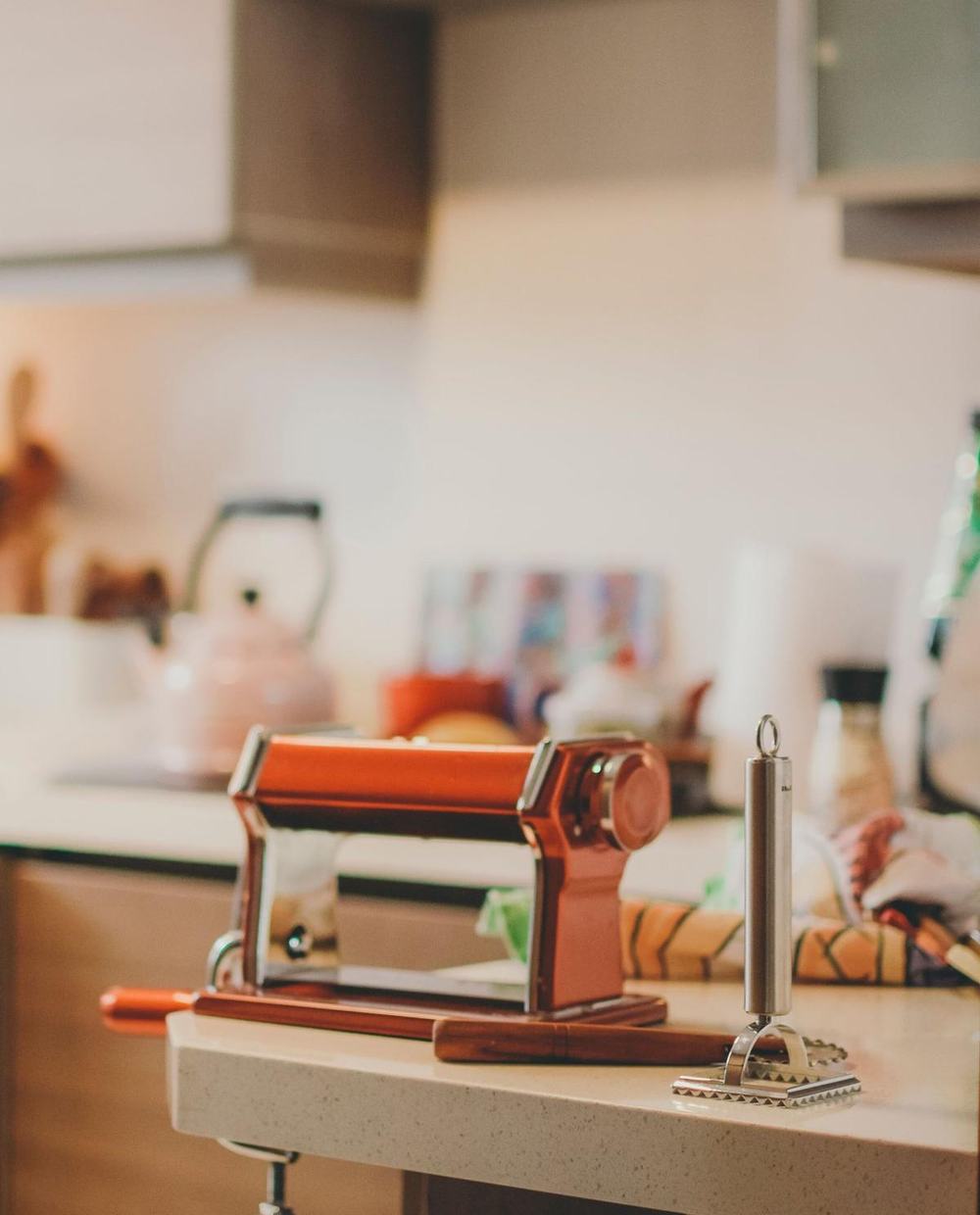 A hand-crank pasta maker clamped to a kitchen counter next to a ravioli stamp