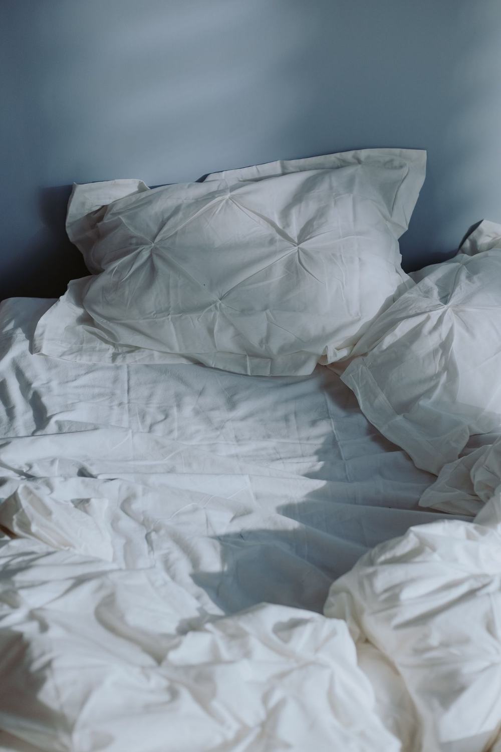 Rumpled white pillows and cotton bedding against a grey-blue wall