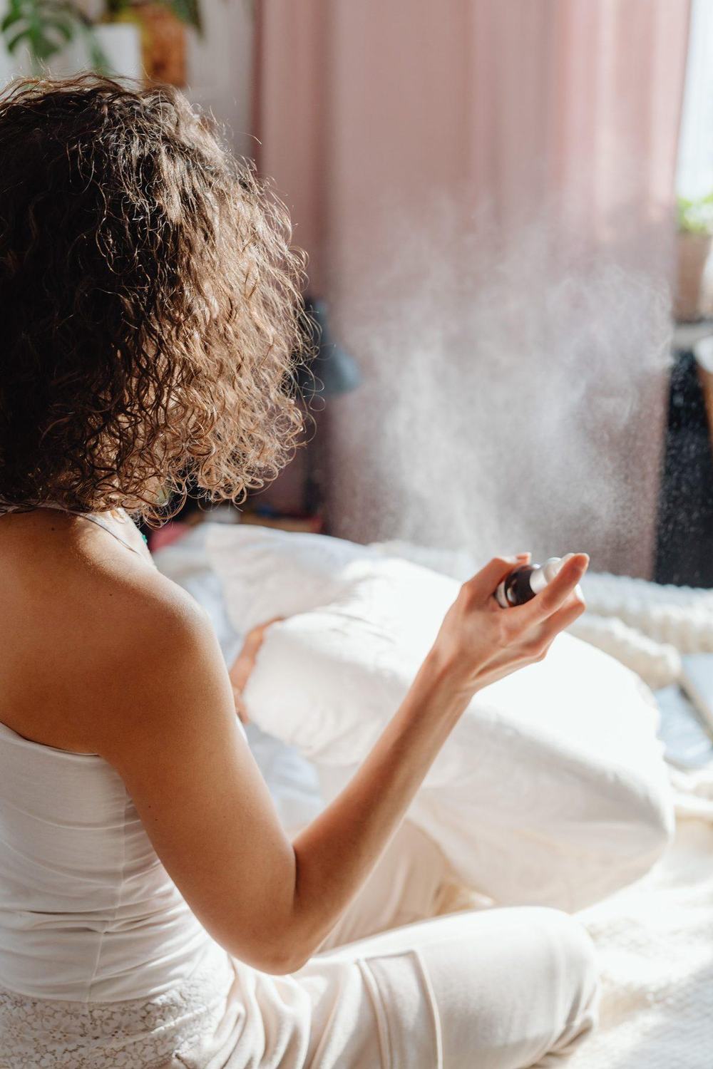Woman with curly hair spraying pillow spray over white bedding in a sunlit bedroom