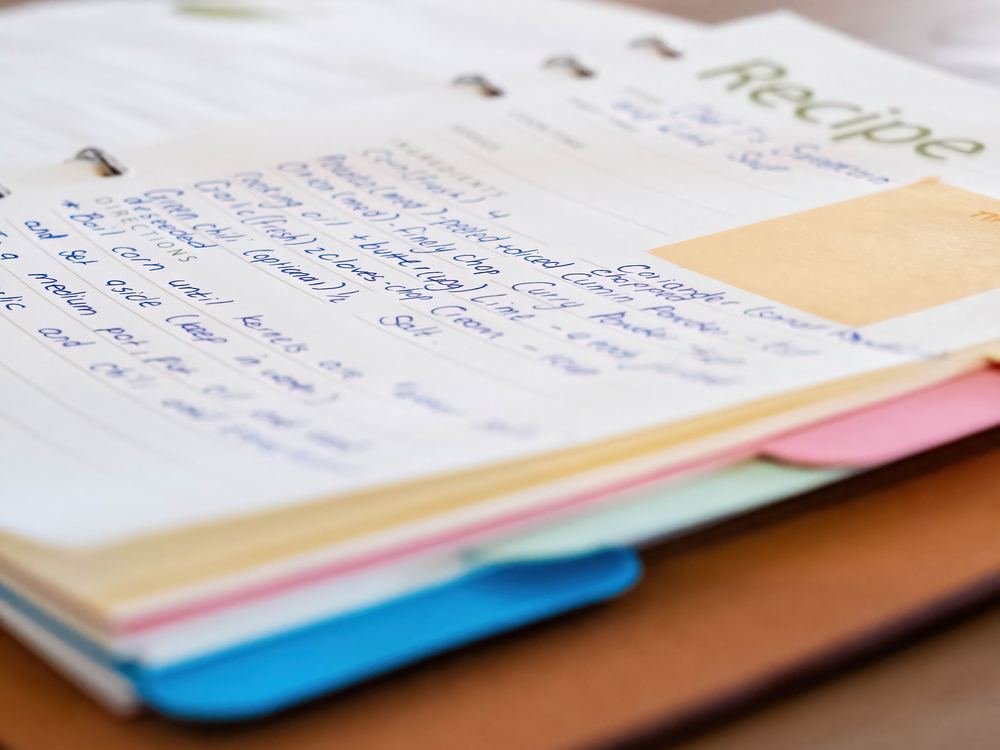 Handwritten recipe pages and colourful folders stacked on a wooden kitchen table