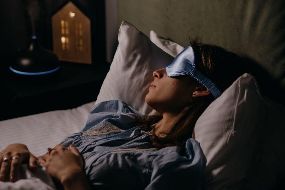 Woman resting in bed wearing a light blue silk sleep mask in a dark bedroom