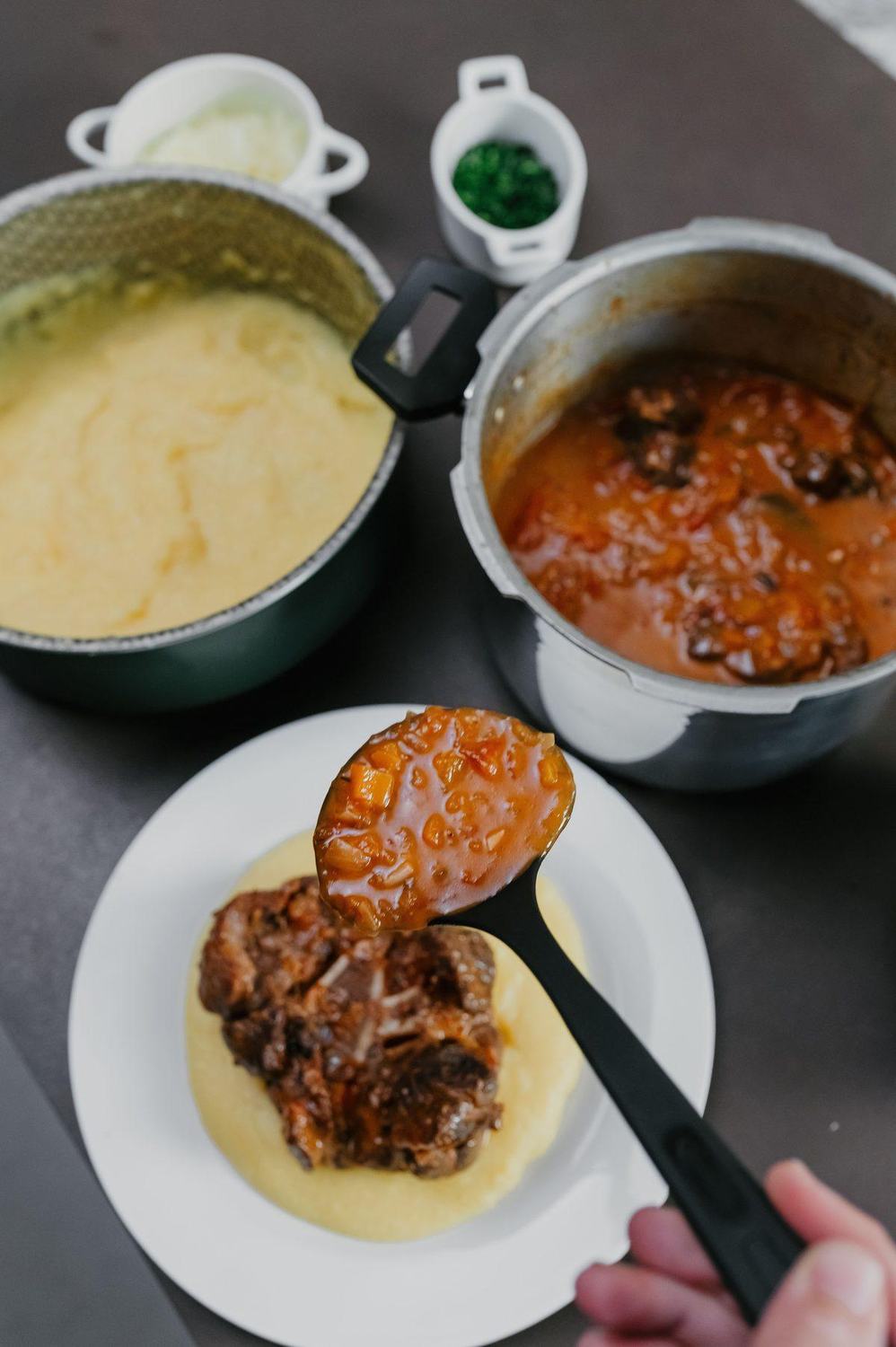 Braised beef stew over creamy polenta, with sauce on a ladle and pots behind