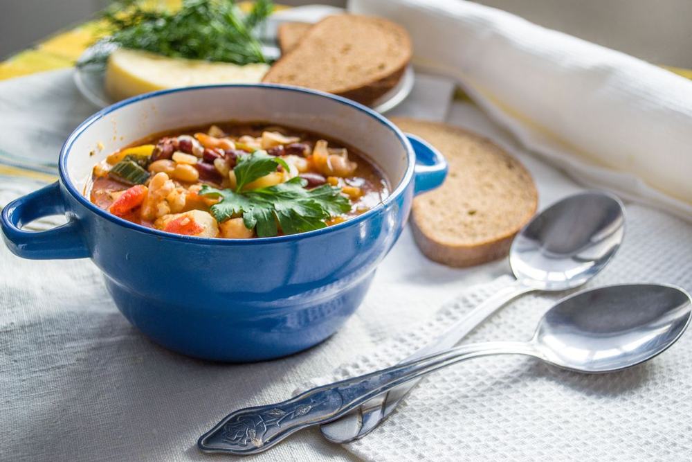 A blue ceramic bowl with handles filled with vegetable bean soup, with bread and spoons