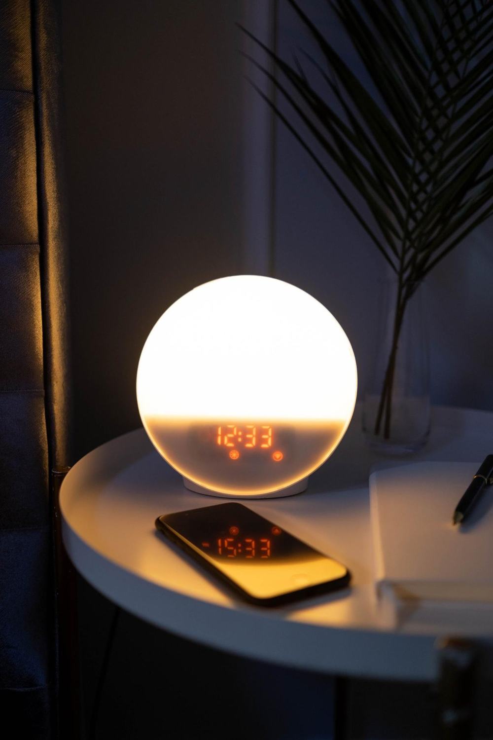 A round sunrise alarm clock glowing warm white on a bedside table in a dark room