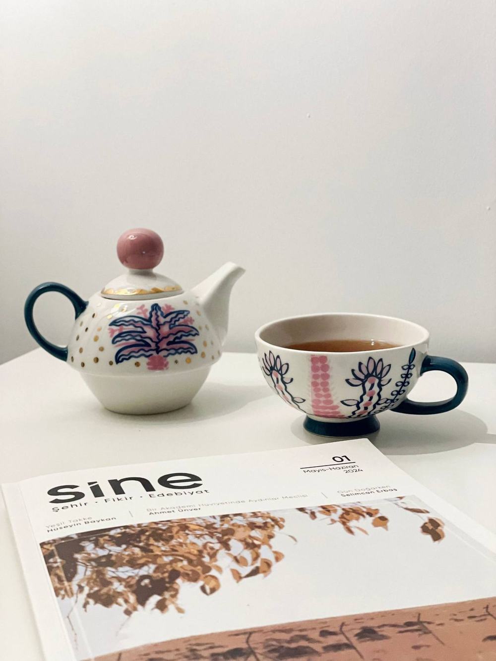A hand-painted tea for one teapot and a cup of tea on a white table beside a magazine