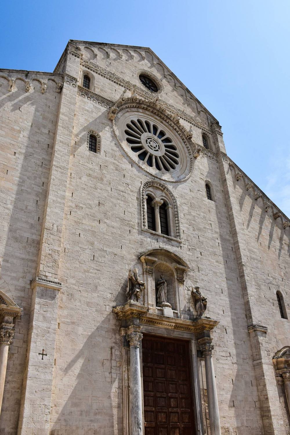 The pale stone Romanesque facade of a cathedral in Bari, with a large rose window above a heavy wooden door