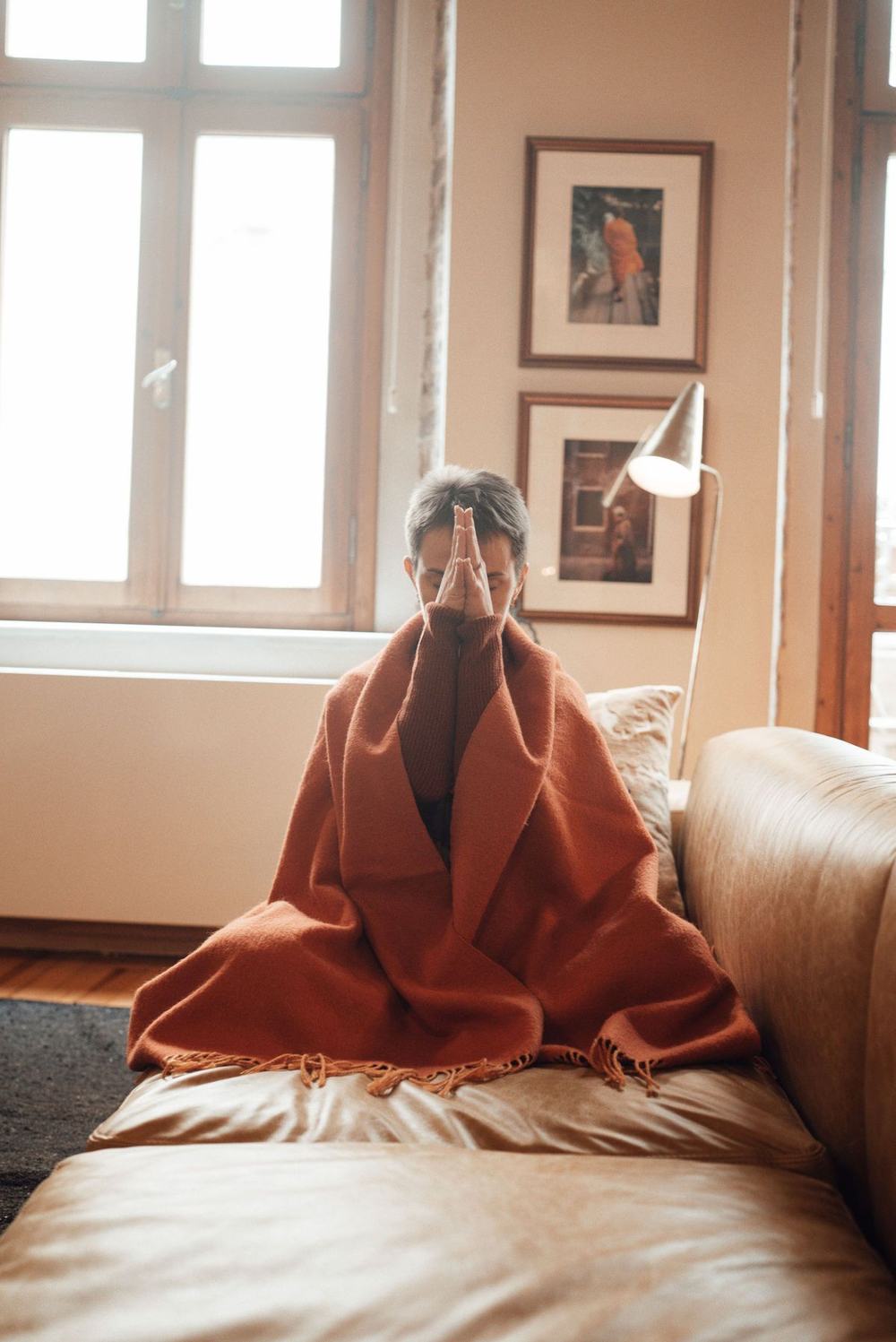 A woman wrapped in a rust-colored blanket on a leather sofa in a sunlit room
