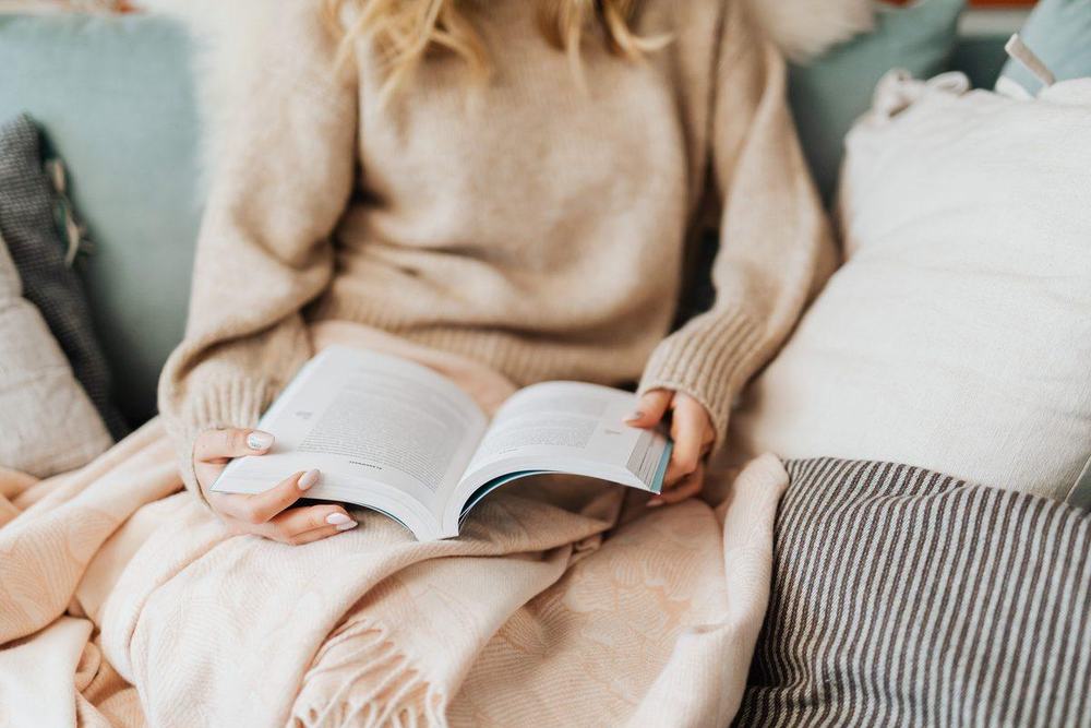 Woman in a beige sweater reading on the sofa under a soft peach blanket