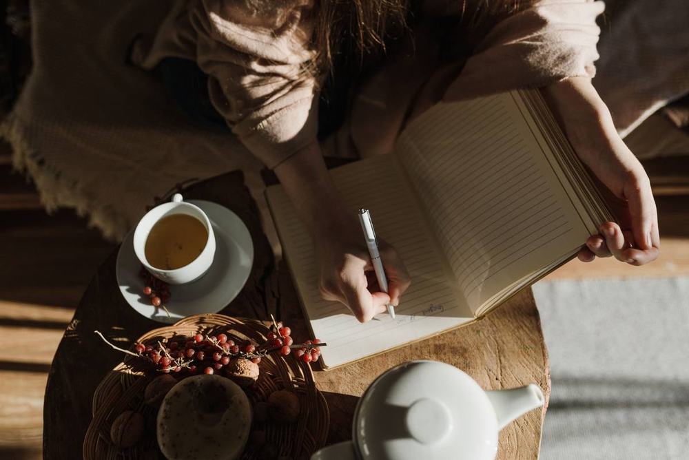 Hands writing in a notebook beside a cup of tea in soft morning light